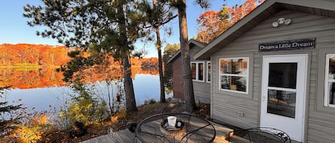 A cabin with a deck, outdoor seating, and a view of a lake and autumn foliage.