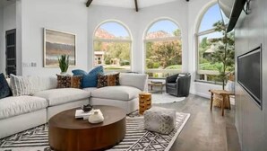 A modern living room with a sectional sofa, a coffee table, and a TV. There are large windows with a view of a garden and mountains. - Chapel Showstopper! Walk to trails, Private yard (Sedona)