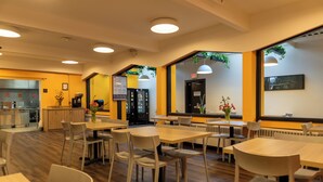 A modern dining area with wooden tables and chairs, a kitchen in the background, and large windows with plants. - HI Vancouver Downtown - Hostel (Vancouver)
