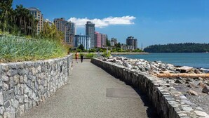 A coastal walkway with a stone wall, a clear view of the sea, and high-rise buildings in the background. - West Vancouver Ambelside Mid-Century Home (West Vancouver)
