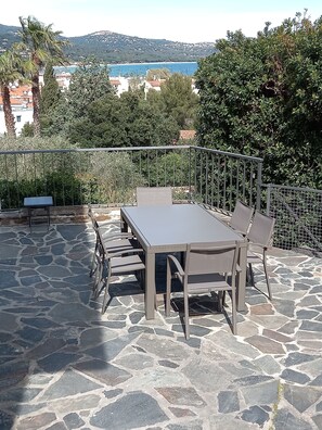 An outdoor patio with a stone floor, a table with chairs, and a view of the sea and hills in the background. - LARGE APARTMENT WITH SEA VIEW (Cavalaire-sur-Mer)