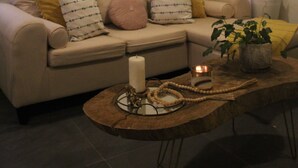 Een woonkamer met een beige bank, gele en witte kussens, een ronde salontafel met een kaars en een klein plantje, en een wandplank met ingelijste foto’s en een hangend decoratieobject.