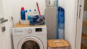 Un área de lavandería compacta con lavadora de carga frontal, una cesta de ropa de madera y un caja de almacenamiento etiquetada como 'LIGHTS' y 'DARKS'.
