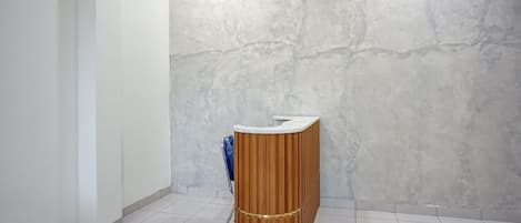 A reception desk with a wooden finish and a white top, situated in a hallway with a tiled floor and a textured wall.