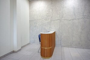 Un mostrador de recepción con detalles de madera y una superficie blanca, ubicado en un pasillo con suelo de baldosas y pared texturizada.