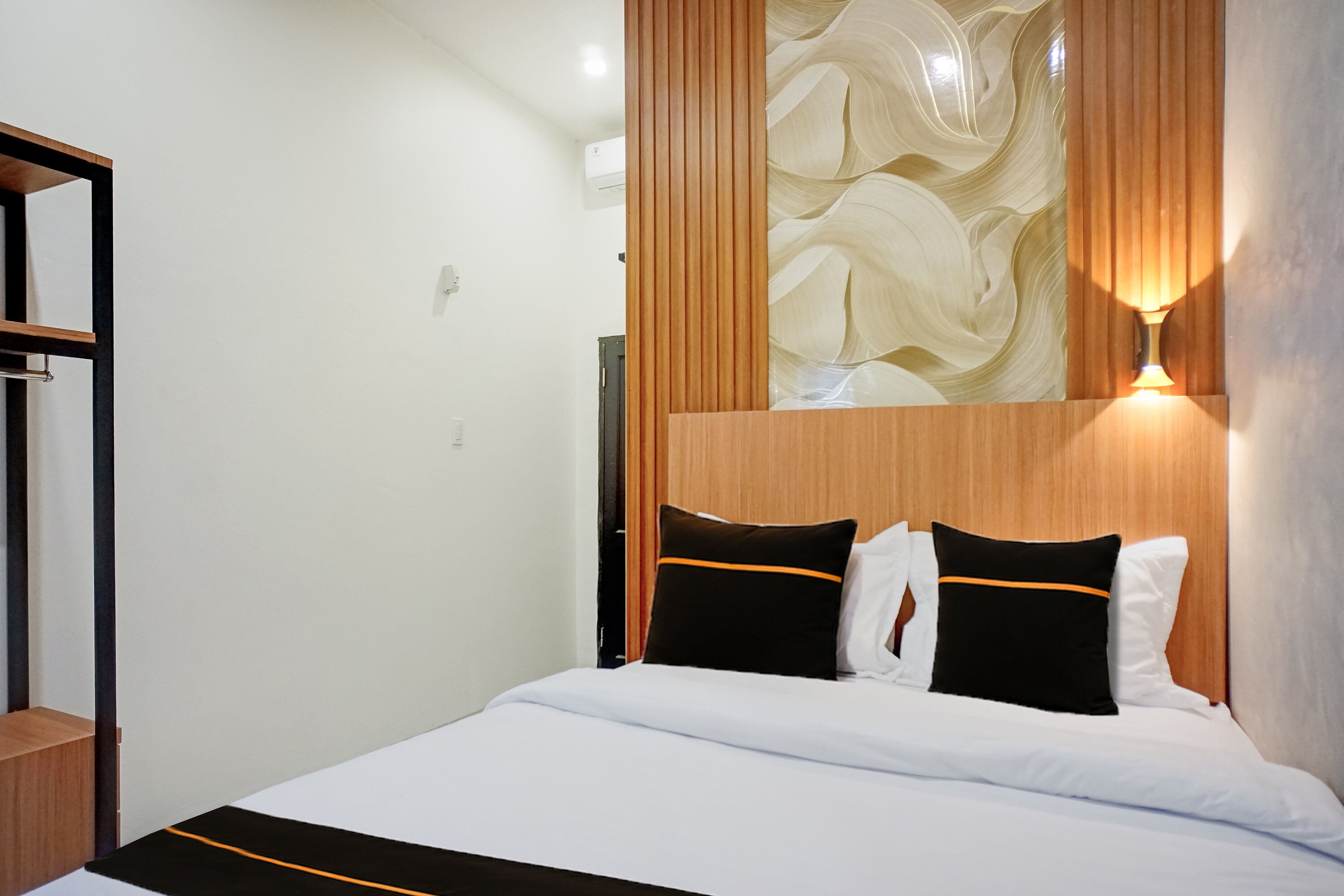 A hotel room with a bed, a nightstand, a mirror, a wall-mounted light, and a decorative panel.