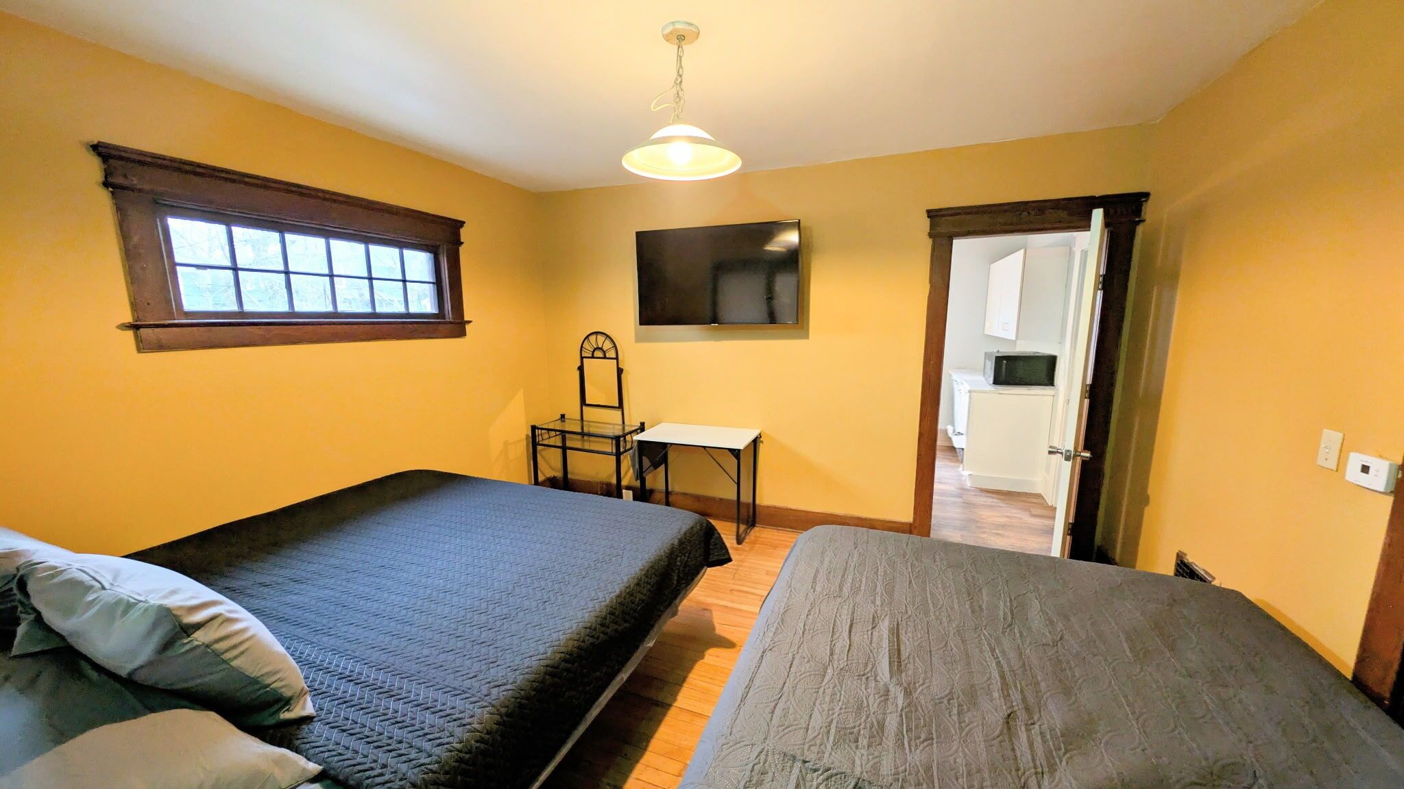 A room with two beds, a desk, a chair, a television, and a door leading to a kitchen.