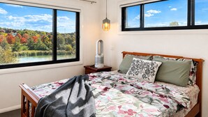 A bedroom with a bed, a nightstand, a lamp, and two windows with a view of a lake and autumn trees.