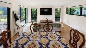 A dining area with a table set for four, a television mounted on the wall, and large windows offering views of greenery.