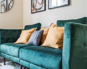 A teal velvet sofa with three cushions, a patterned pillow, and a framed abstract painting on the wall. - Homely Stay | Fast Wi-Fi|Smart TV|Balcony (Birmingham)
