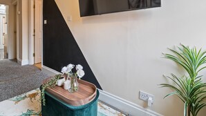 Un corridoio con parete in bianco e nero, un cartello Netflix, un tavolino in legno con un vaso di fiori bianchi e una pianta verde.