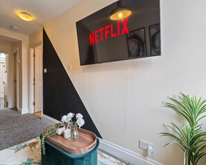 A hallway with a black and white wall, a Netflix sign, a wooden side table with a vase of white flowers, and a green plant. - Homely Stay | Fast Wi-Fi|Smart TV|Balcony (Birmingham)