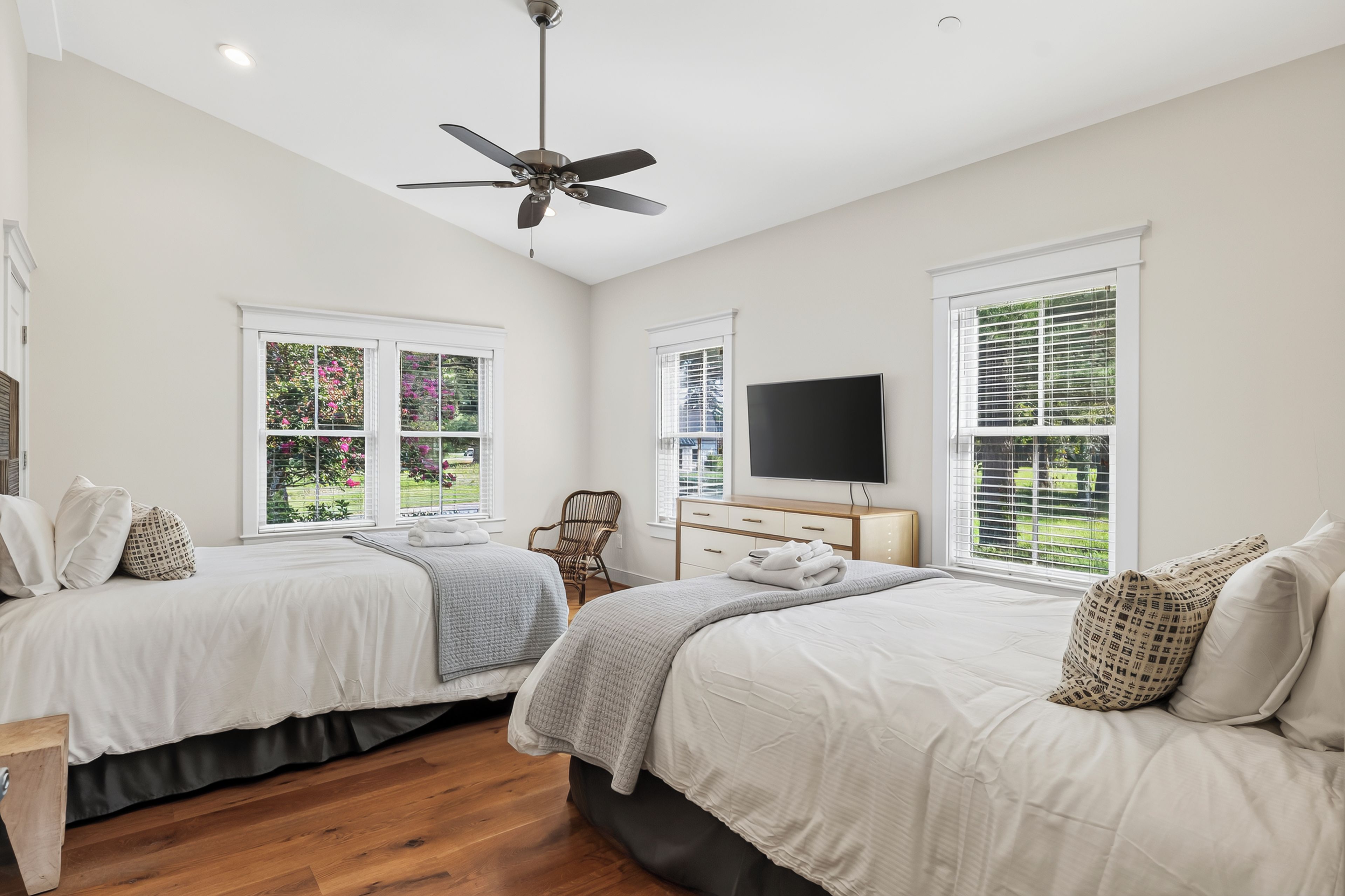 A bedroom with two beds, a ceiling fan, a TV, and large windows with blinds.