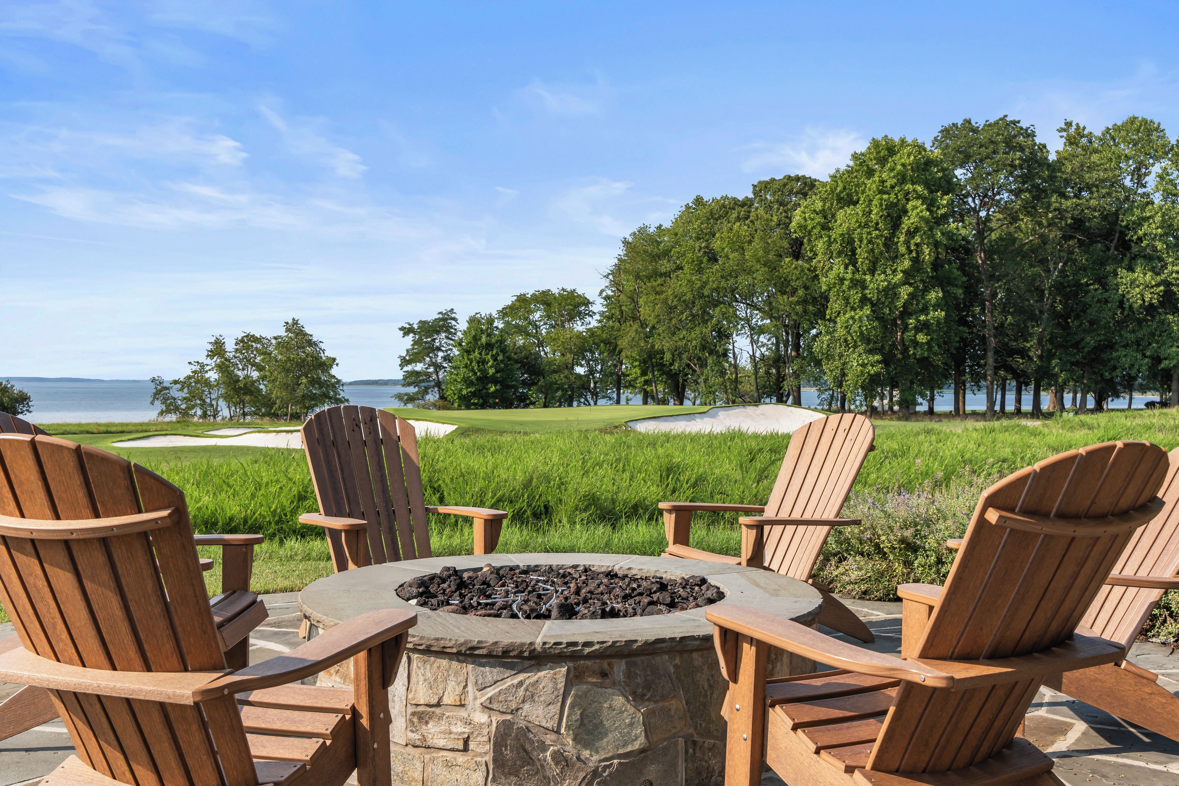 A stone fire pit surrounded by wooden Adirondack chairs, overlooking a golf course and a body of water.