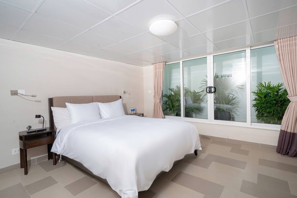 A hotel room with a large bed, a nightstand, a lamp, a window with curtains, and a view of greenery outside.
