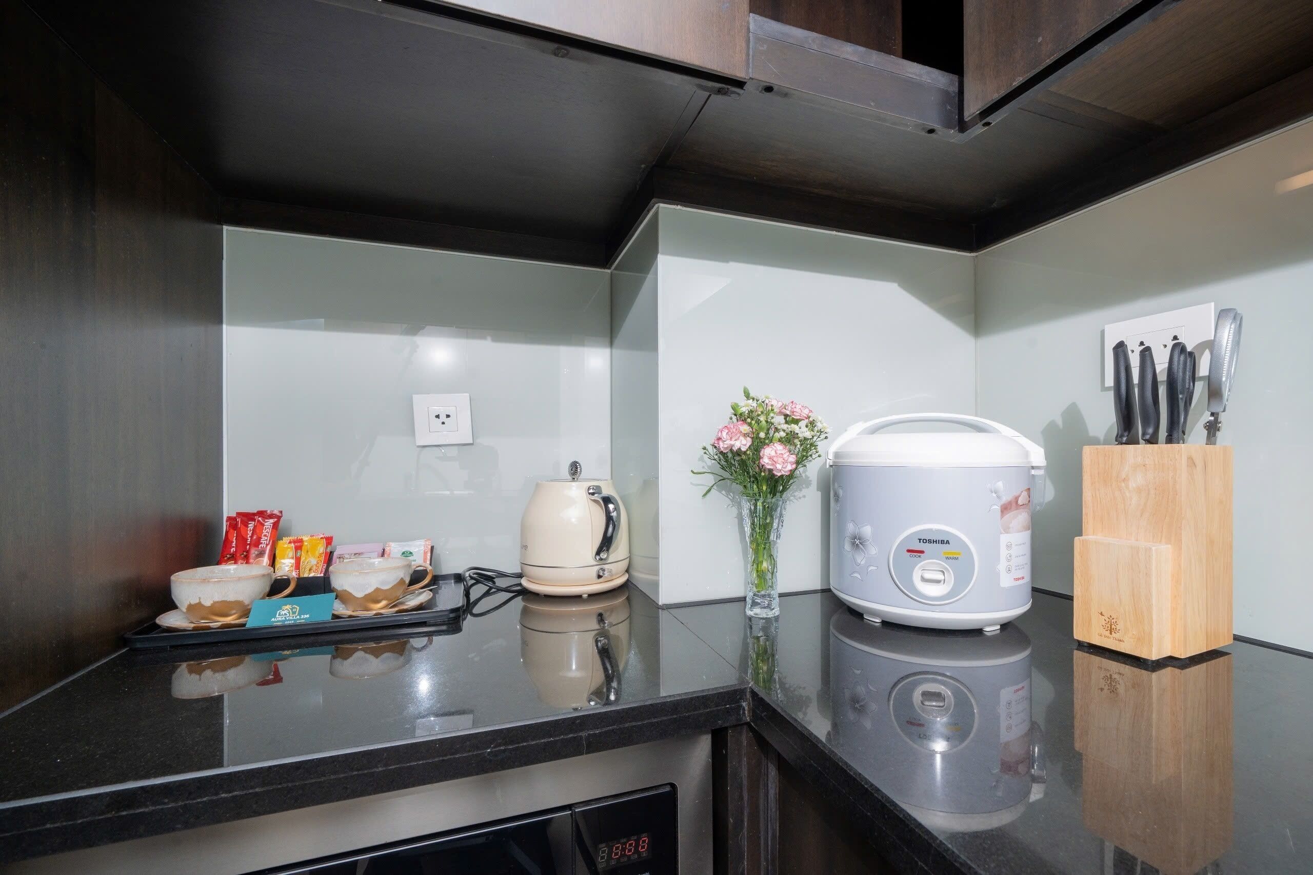 Une cuisine avec un plan de travail noir, un cuiseur à riz blanc, une machine à café et un vase rempli de fleurs.