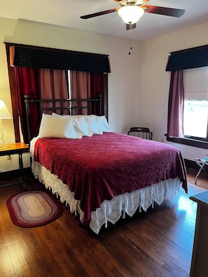 A neatly made bed with a red bedspread and white pillows, a wooden floor, a ceiling fan, and a window with curtains.