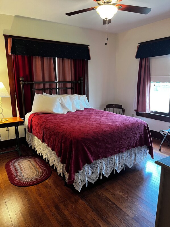 A neatly made bed with a red bedspread and white pillows, a wooden floor, a ceiling fan, and a window with curtains.