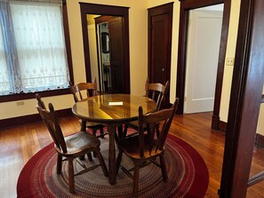 A dining area with a round glass table surrounded by wooden chairs, a red woven rug, and a window with patterned curtains.