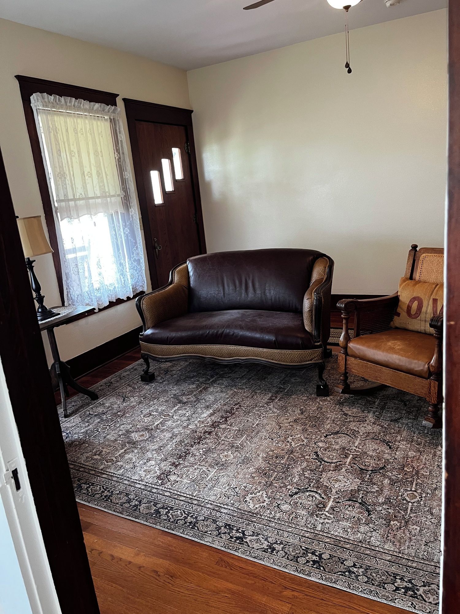 A room with a dark brown leather sofa, a patterned rug, a small wooden table, and a window with sheer curtains.
