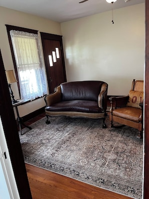 A room with a dark brown leather sofa, a patterned rug, a small wooden table, and a window with sheer curtains.