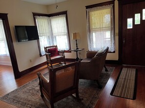 A living room with a wooden floor, a sofa, a chair, a small table with a lamp, and a television mounted on the wall.