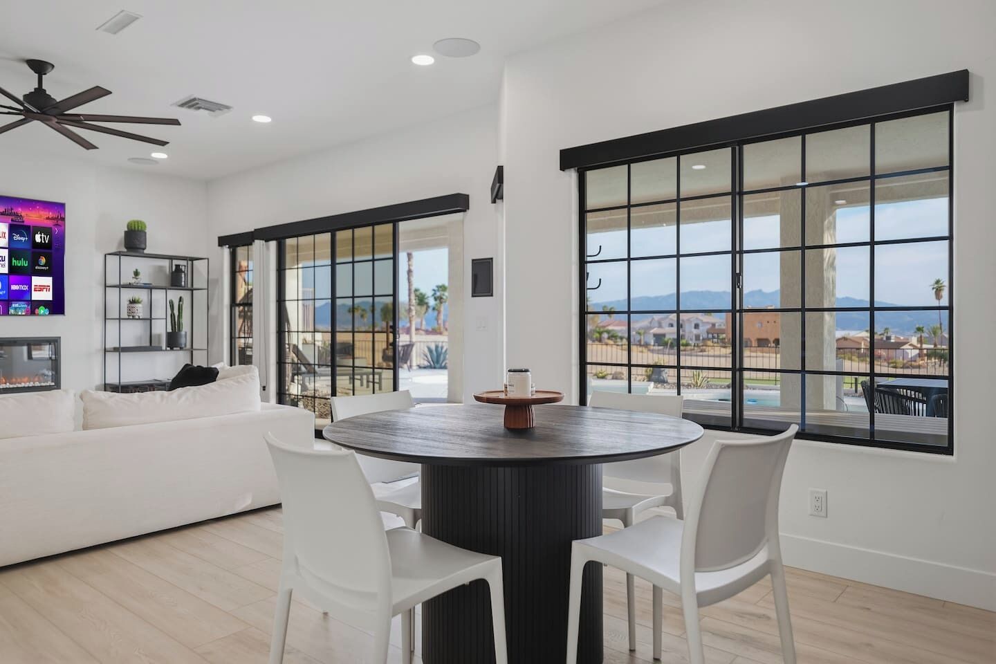 A modern living room with a round dining table, white chairs, a white sofa, and large windows with a view of a residential area.