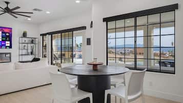 A modern living room with a round dining table, white chairs, a white sofa, and large windows with a view of a residential area.