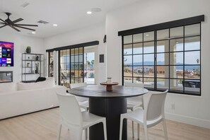Ein modernes Wohnzimmer mit einem runden Esstisch, weißen Stühlen, einem weißen Sofa und großen Fenstern mit Blick auf eine Wohngegend.