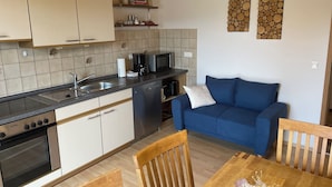 Een kleine keuken met een vaatwasser, kookplaat en magnetron. In de woonruimte bevindt zich een blauwe bank en houten stoelen.