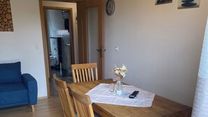 Een eetruimte met een houten tafel, stoelen en een vaas met bloemen. Achtergrond is een blauwe bank.
