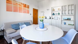Ein modernes Wohnzimmer mit einem runden weißen Tisch, blauen Stühlen, einem gestreiften Sofa und einer weißen Schrankwand mit Regalen.