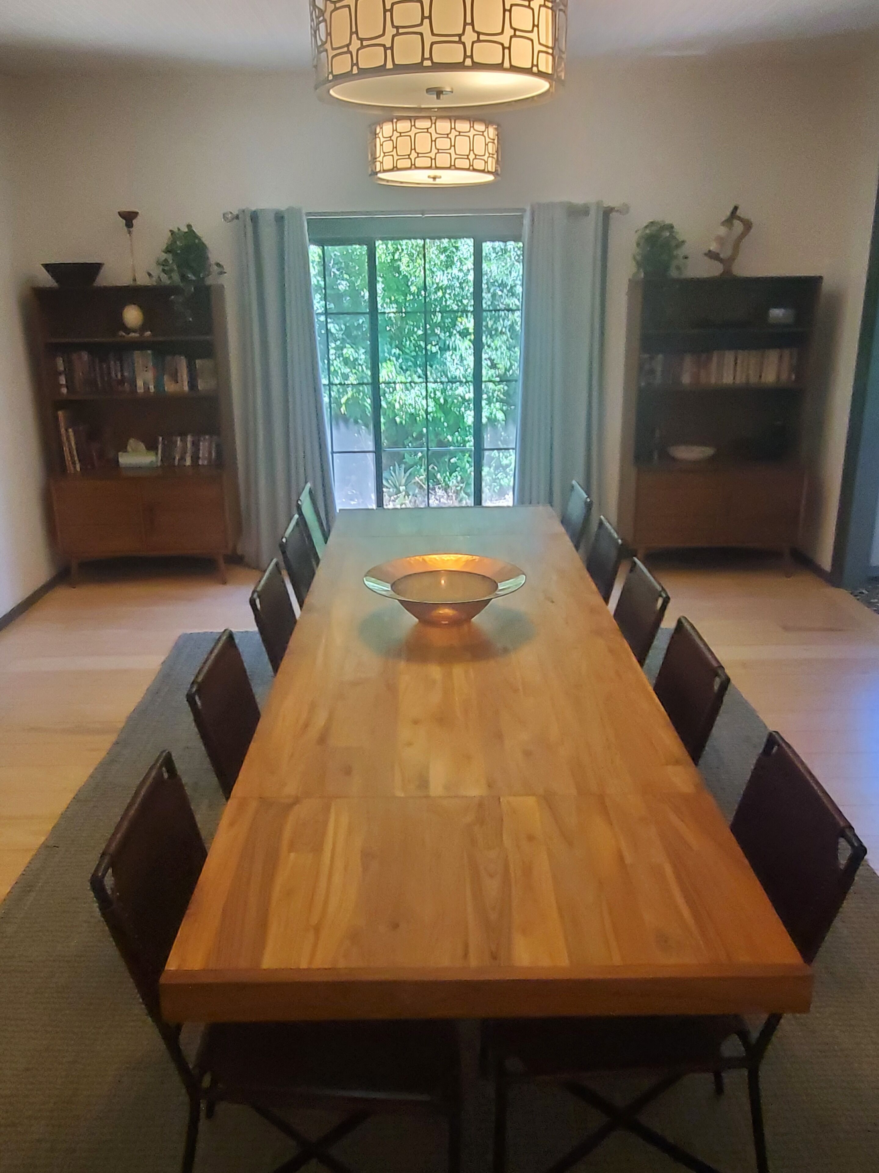 Une salle à manger avec une longue table en bois, des chaises, un luminaire central et des étagères remplies de plantes.