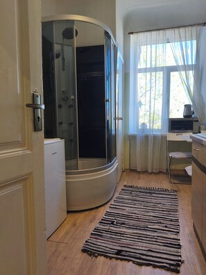 A room with a curved shower enclosure, a microwave, a small refrigerator, and a window with curtains. - Sunny Studio Apt with Free Parking (Rīga)