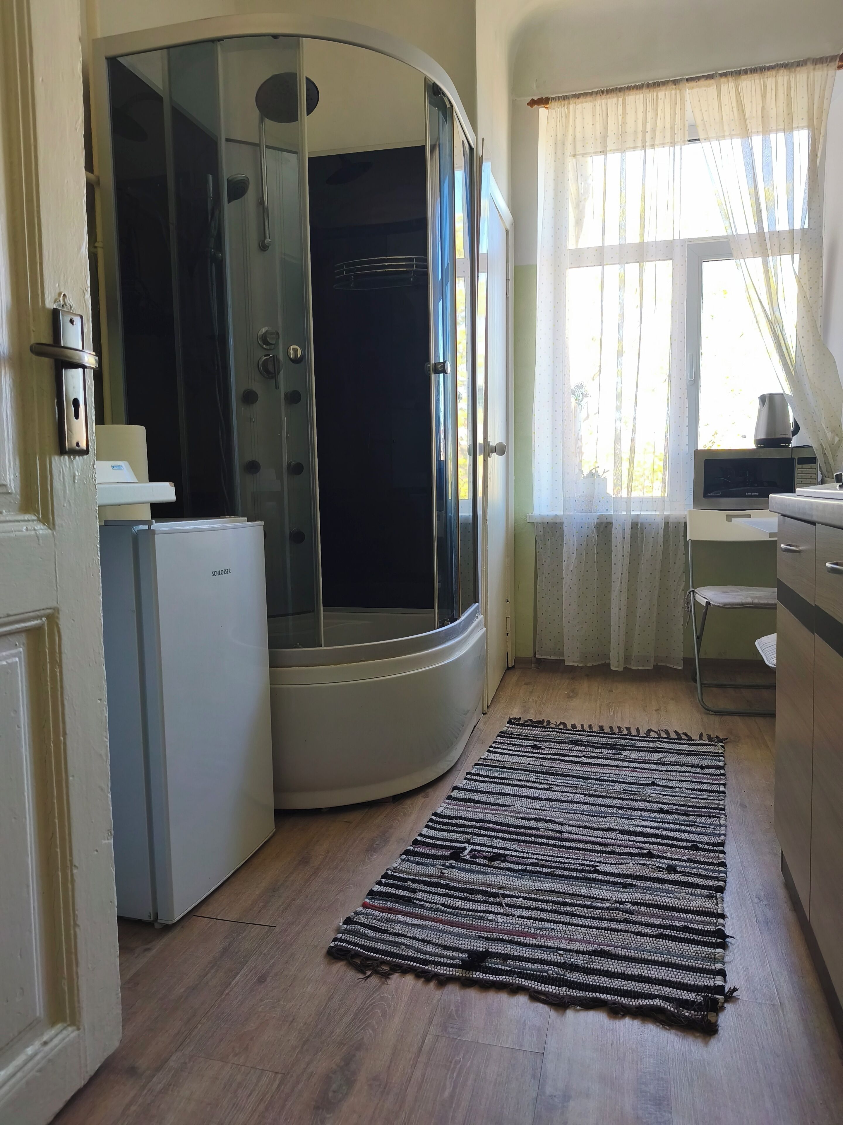 A compact room with a shower, a refrigerator, a microwave, a small table, and a window with curtains.