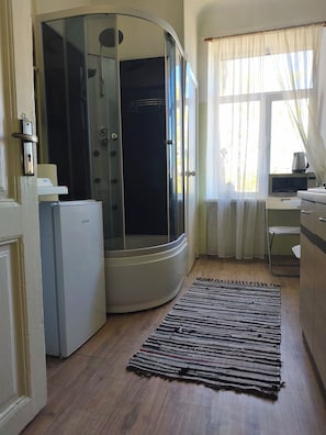 A compact room with a shower, a refrigerator, a microwave, a small table, and a window with curtains. - Sunny Studio Apt with Free Parking (Rīga)