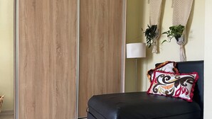 Una habitación con un armario corredero de madera, un sofá de cuero negro, un cojín estampado, una lámpara de pie y adornos de macramé en las paredes.