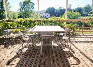 Eine Holzterrasse mit Tisch und Stühlen, umgeben von Grünpflanzen und einem Haus im Hintergrund.