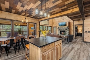 A rustic cabin interior with a large dining area, kitchen, and living space, featuring wooden floors and a stone fireplace. - Bluff Ridge Retreat + SwimSpa + Hot Tub+ Air Hockey + (Sevierville)