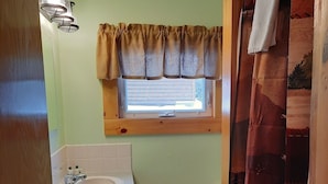Ein Badezimmer mit Holzwaschtisch, einem Fenster mit Vorhang und einer Duschkabine mit Waldmusterung.