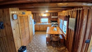 Interior d'una cabana amb parets de fusta, una cuina sencera, una taula de dinar i un cub de brossa.