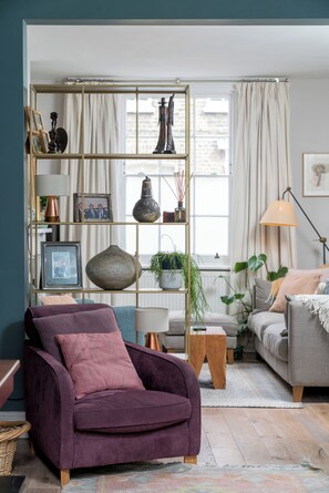 Ein Wohnzimmer mit einem violetten Sessel, einem grauen Sofa, einem Holztisch und einem Bücherregal mit verschiedenen Dekorationsobjekten.