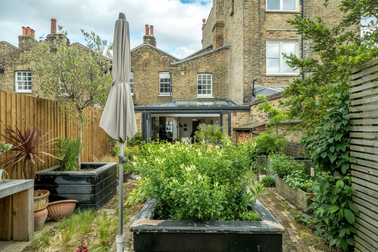 A garden with a patio area, a wooden fence, and a brick building in the background.