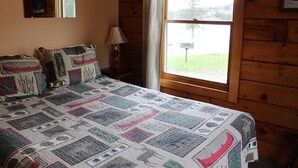 Quarto com cama, luminária, janela com vista para área gramada e paredes de madeira.