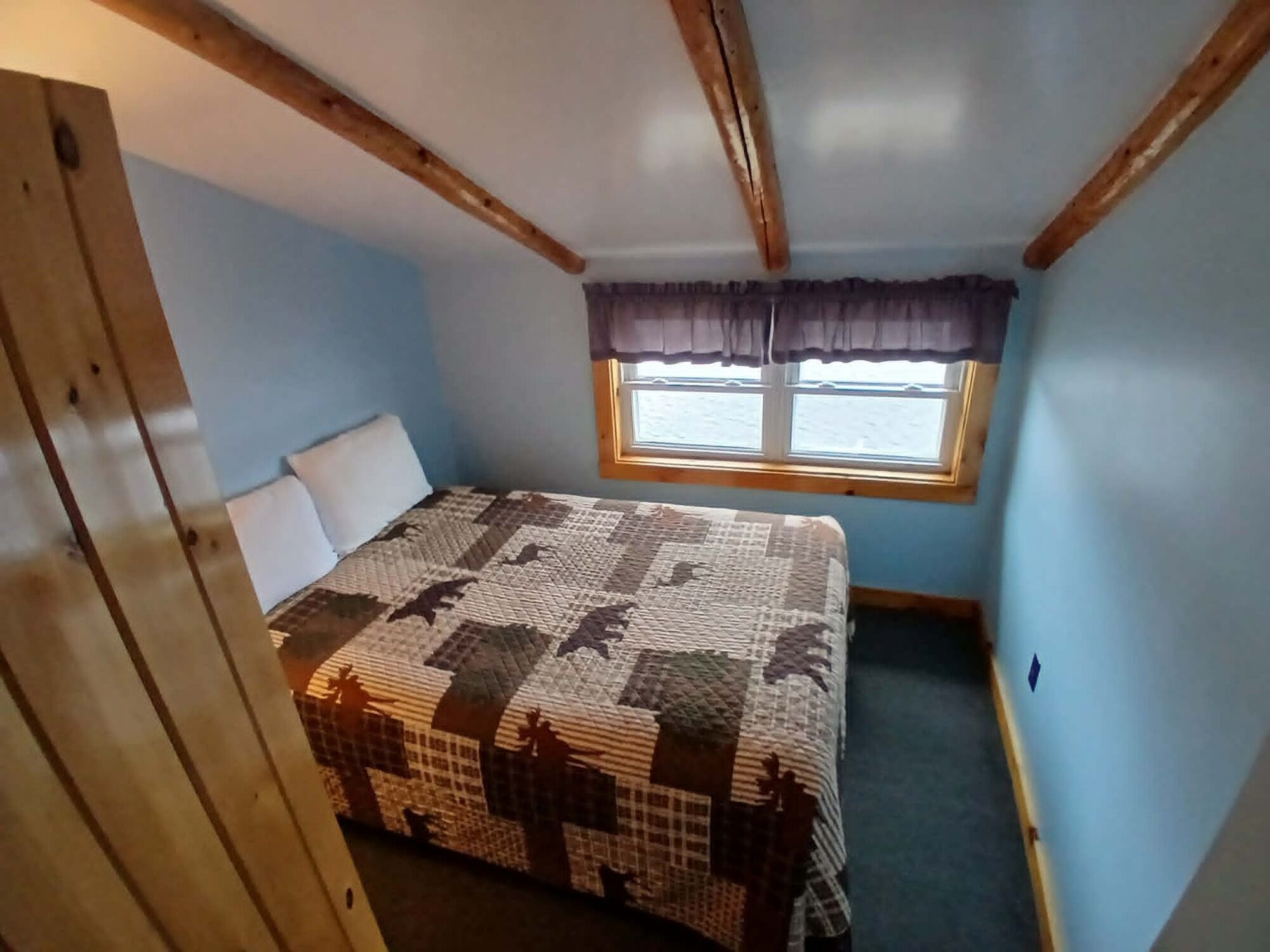 Un dormitorio con cama, ventana con cortinas y vigas de madera a la vista.