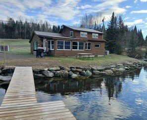 Una baita di legno con un molo che si protende su un lago, circondata da un muro di pietra e un'area verde.