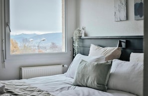 A bedroom with a bed, a window, a radiator, and a wall-mounted shelf with a vase. - Les Loges Du Leman (Crozet)