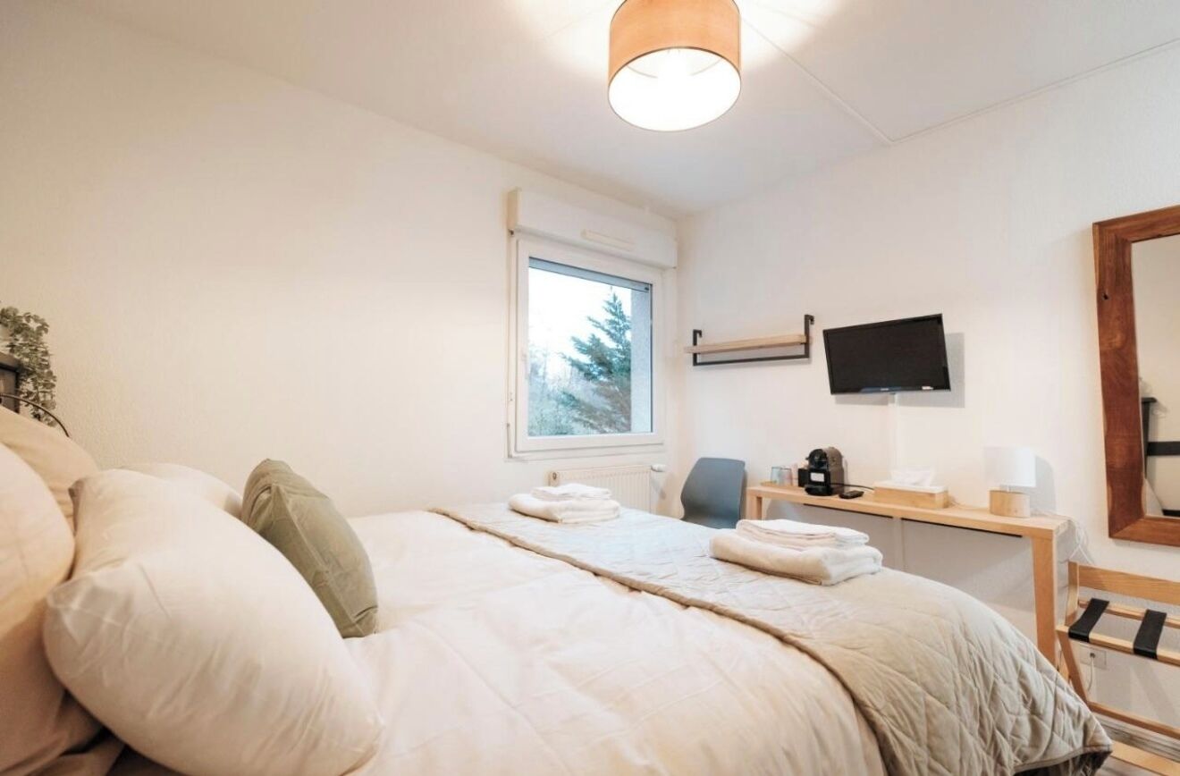 Ett hotellrum med en säng, ett skrivbord med en dator, en tv och ett fönster med utsikt över träd.