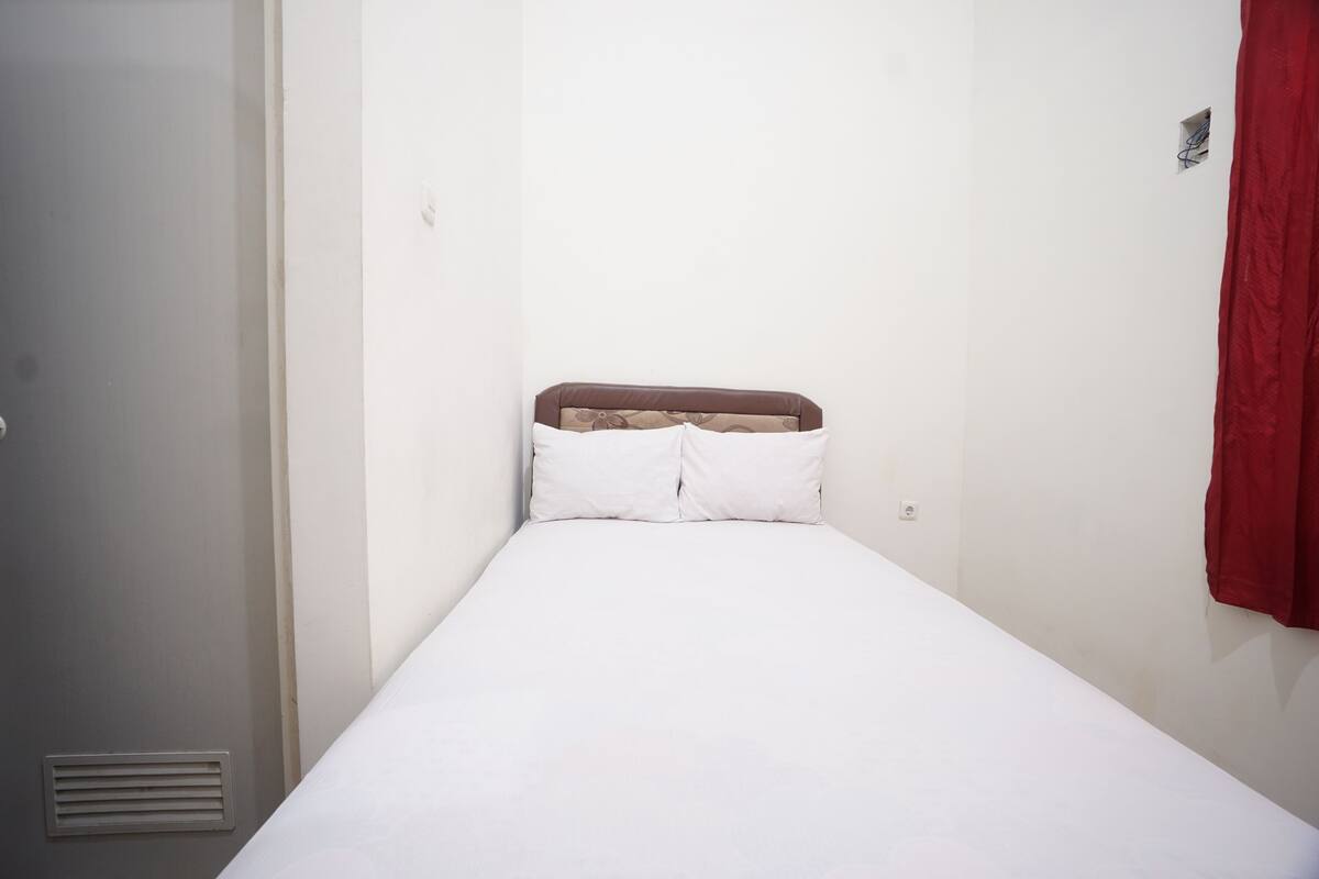 A small, single-bed room with a plain white wall, a brown headboard, and a red curtain.