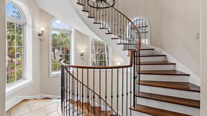 Una imponente escalera de madera con barandilla y balaustres negros, que conduce a un rellano con amplias ventanas y un candelabro.